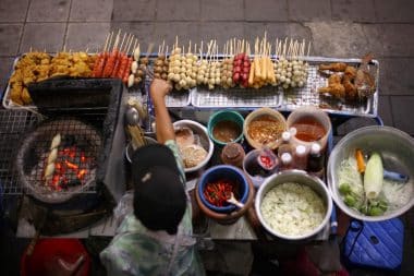 Ein Straßenverkäufer in Bangkok bereitet auf einem Holzkohlegrill Spieße und frittierte Snacks zu, umgeben von Schüsseln mit Soßen, Gemüse und Gewürzen auf dem belebten Bürgersteig eines Marktes.