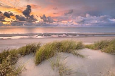 Im Vordergrund blickt man über das Dünengras auf einen Sandstrand und das ruhige Meer bei Sonnenuntergang, während bunte Wolken den Himmel füllen – eine klassische Küstenlandschaft in den Niederlanden.