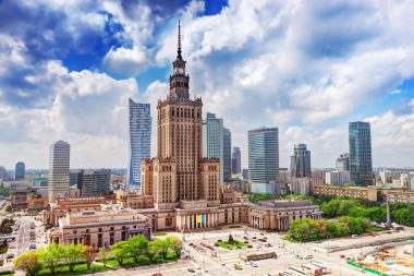 Inmitten einer polnischen Stadtlandschaft mit modernen Wolkenkratzern steht unter einem teilweise bewölkten Himmel ein hohes, kunstvoll verziertes Gebäude mit einer Turmspitze. Grüne Bäume und städtische Straßen umgeben die Gebäude.
