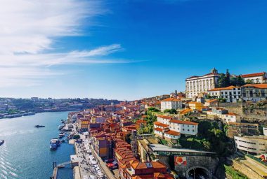 Blick auf das Flussufer von Porto, Portugal, mit bunten Häusern, Booten auf dem Douro und historischen Gebäuden an einem sonnigen Tag unter blauem Himmel – ein Bild, das den lebendigen Charme dieser ikonischen Stadt Portugals einfängt.