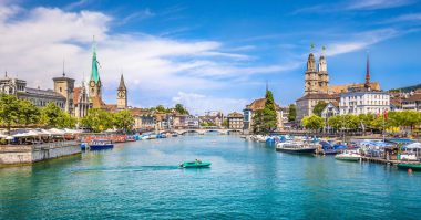 Ein malerischer Blick auf Zürich, Schweiz, mit einem ruhig fließenden Fluss mit Booten, historischen Gebäuden, Kirchtürmen und einer Steinbrücke unter strahlend blauem Himmel mit vereinzelten Wolken.