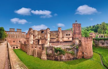 Ein großes, historisches Steinschloss, das an Heidelberg erinnert, steht mit teilweise verfallenen Mauern auf einer grünen Wiese unter einem strahlend blauen Himmel mit vereinzelten Wolken. Im Hintergrund sind Bäume und bewaldete Hügel zu sehen.