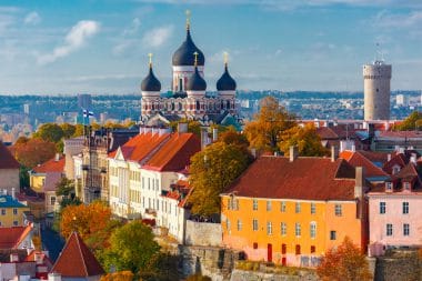 Farbenfrohe alte Gebäude und Bäume in Tallinn, Estland, mit den Zwiebeltürmen der Alexander-Nevski-Kathedrale und einem Steinturm, die im Hintergrund unter blauem Himmel zu sehen sind und den Charme der historischen Skyline von Tallinn unterstreichen.