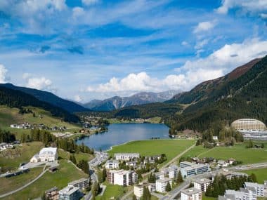 Luftaufnahme des Davoser Sees in der Schweiz, umgeben von grünen Bergen, Kiefernwäldern und den Gebäuden der Stadt.