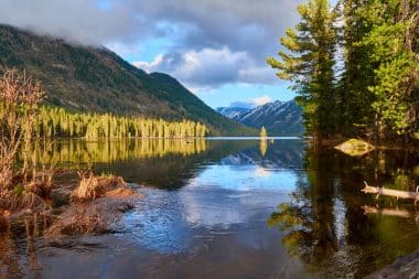 Ein ruhiger Bergsee in Kasachstan spiegelt unter einem teilweise bewölkten Himmel Kiefern und schneebedeckte Gipfel wider, während im Vordergrund sanft Wasser fließt.
