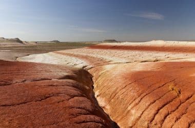 Bunte, geschichtete Wüstenlandschaft in Kasachstan mit roten, orangefarbenen und weißen Schichten unter blauem Himmel; am Horizont sind flache Hügel zu sehen.