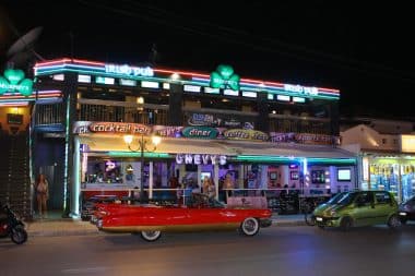 Vor einer belebten, neonbeleuchteten Bar mit Restaurant in Laganas steht nachts ein knallrotes Oldtimer-Cabrio; drinnen sitzen Gäste, und bunte Schilder werben für einen Irish Pub, ein Café, eine Sportbar und Cocktails.