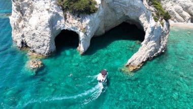 Ein kleines Boot fährt in der Nähe von Meereshöhlen, die sich in die weißen Klippen über dem klaren, türkisfarbenen Wasser von Laganas eingegraben haben, während in der Nähe ein Schwimmer zu sehen ist.