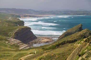 Felsige Klippen und grüne Hügel überragen die zerklüftete Küste Kantabriens, wo sich die Wellen an einem Sandstrand brechen. Das Meer schimmert türkis, der Himmel ist teilweise bewölkt, und am Horizont ist in der Ferne Land zu sehen.