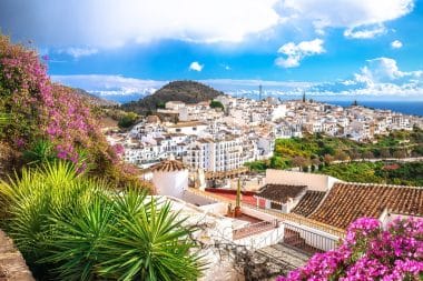 Die weißen Häuser des Bergdorfs Frigiliana blicken auf das ferne Meer, eingerahmt von leuchtend magentafarbenen Bougainvilleen und grünen Pflanzen.