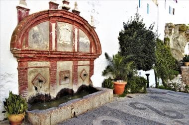 Ein historischer Steinbrunnen mit drei Ausläufen in Frigiliana, der vor einer weißen Wand neben Topfpflanzen und einem gepflasterten Weg steht.