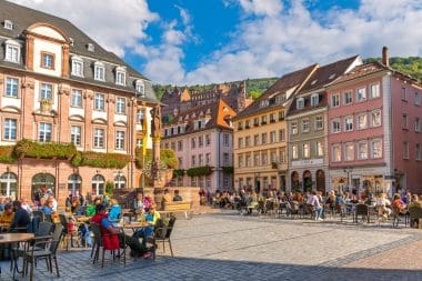 Auf einem belebten europäischen Stadtplatz in Heidelberg sitzen Menschen an Tischen im Freien, umgeben von farbenfrohen historischen Gebäuden unter einem blauen Himmel mit vereinzelten Wolken. Ein Springbrunnen und Blumen schmücken den Platz, und im Hintergrund sind Bäume zu sehen.