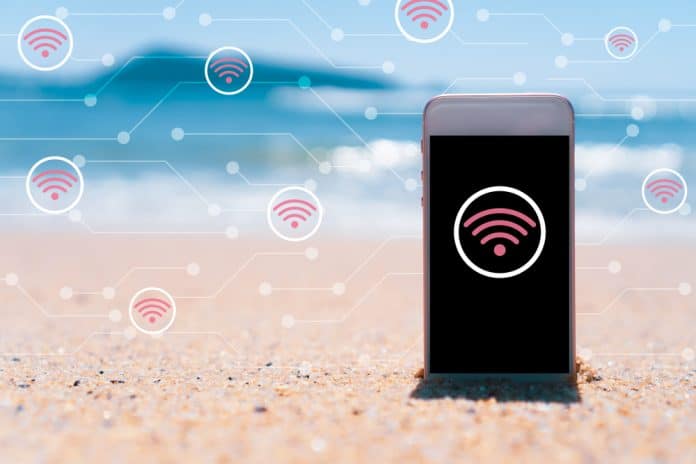 Smart,Phone,On,Tropical,Beach,Blue,Sky,With,Wifi,Icon Handy liegt am Strand mit WLAN-Symbolen