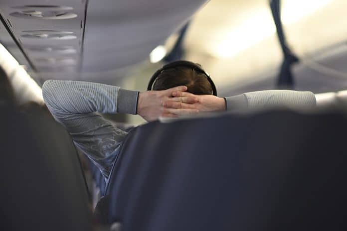 A,Passenger,Relaxing,On,An,Airplane,,Wearing,Headphones,,With,Hands Eine Person mit Kopfhörern sitzt in einem Flugzeug, die Hände hinter dem Kopf verschränkt; Blick von hinten durch die Sitzreihen.