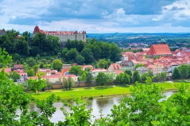 Ein malerischer Blick auf eine europäische Stadt mit Gebäuden mit roten Dächern, einer großen historischen Burg auf einem Hügel, üppig grünen Bäumen und einem Fluss im Vordergrund unter einem bewölkten blauen Himmel.