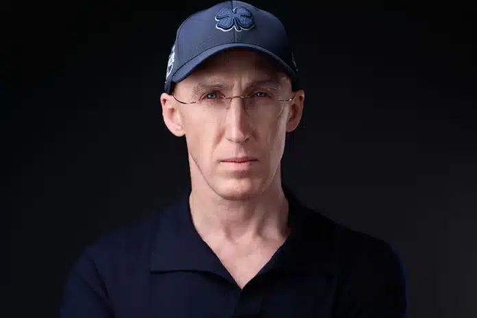 1 Ein Mann mit einer marineblauen Mütze mit einem vierblättrigen Kleeblatt-Emblem und einer Brille, der ein dunkles Hemd mit Kragen trägt, blickt vor einem schwarzen Hintergrund direkt in die Kamera.