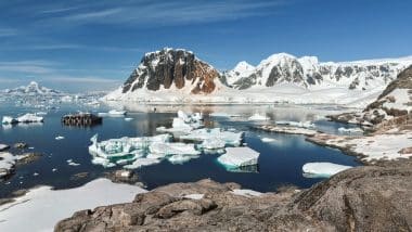 Verschneite Berge und Gletscher umgeben eine ruhige, eisige Bucht mit schwimmenden Eisbergen. Ein Holzsteg ragt ins Wasser hinein, und der Himmel ist klar und blau.