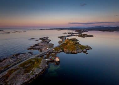 Eine kurvenreiche Küstenstraße, die als eine der schönsten Panoramastraßen Europas gilt, schlängelt sich bei Sonnenuntergang an felsigen Inselchen vorbei, die von ruhigem Wasser umgeben sind; am Ufer steht ein kleines Haus, und in der Ferne sind Berge zu sehen.