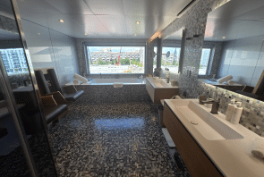Modernes Badezimmer mit Mosaikfliesenboden, zwei Waschbecken, einer Badewanne neben einem großen Fenster mit Blick auf die Stadt und Liegestühlen neben der Badewanne. Handtücher und Pflegeprodukte sind im gesamten Raum ordentlich angeordnet.