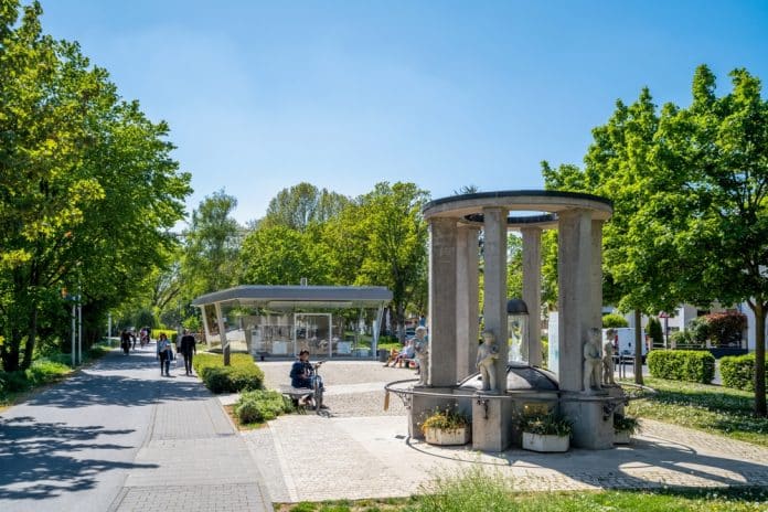 Old,City,Of,Bad,Vilbel,,Hessen,,Germany Ein von Bäumen gesäumter Weg, auf dem Menschen spazieren gehen und Rad fahren, im Vordergrund ein runder Steinbrunnen mit Statuen und im Hintergrund ein modernes Glasgebäude an einem sonnigen Tag.