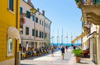 Eine sonnige Straße am Meer, auf der Menschen spazieren gehen und in Straßencafés essen, gesäumt von bunten Gebäuden. Im Hintergrund liegen Segelboote mit Flaggen unter einem strahlend blauen Himmel vor Anker.