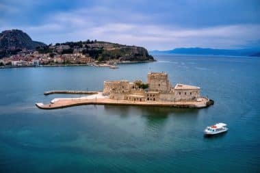Auf einer von blauem Wasser umgebenen Insel thront eine kleine Steinfestung, im Hintergrund sind eine Küstenstadt und Hügel zu sehen, und ein weißes Boot nähert sich der Insel.