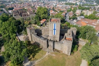 Luftaufnahme einer mittelalterlichen Steinburg, einer der bekanntesten Sehenswürdigkeiten von Guimarães, mit einem großen weiß-blauen vertikalen Banner, umgeben von Bäumen und einer Stadt mit rotgedeckten Gebäuden im Hintergrund.