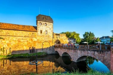 Ein historisches Stein-Schloss mit einer Bogenbrücke, die über einen ruhigen Burggraben führt; unter strahlend blauem Himmel versammeln sich Menschen auf der Brücke und in der Nähe des Schlosseingangs.