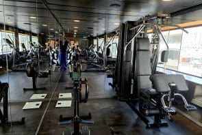 Ein modernes Fitnessstudio mit verspiegelten Wänden, verschiedenen Trainingsgeräten, einem Heimtrainer und zwei Personen, die ein Foto machen. Große Fenster sorgen für Tageslicht.
