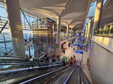 In einem modernen, großzügigen Flughafenterminal, das von Tageslicht durchflutet ist und über große Fenster, hohe Decken sowie digitale Fluginformationsanzeigen verfügt, fahren Menschen die Rolltreppen hinunter. Der Blick fällt auf den Abflugbereich.