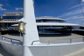 Eine Nahaufnahme des Decks einer modernen Yacht, auf dem eine Messingglocke an einer weißen Konstruktion befestigt ist, vor dem Hintergrund einer eleganten Architektur mit Glaswänden und einem blauen Himmel.