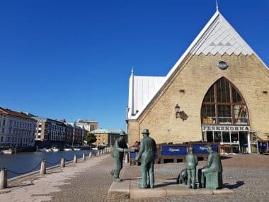 Ein sonniger Tag in der Markthalle Feskekôrka in Göteborg, Schweden, mit bronzenen Fischhändler-Statuen im Vordergrund und Gebäuden am Kanal unter strahlend blauem Himmel – eine perfekte Kulisse, um eine der beliebtesten Sehenswürdigkeiten Göteborgs zu erleben.