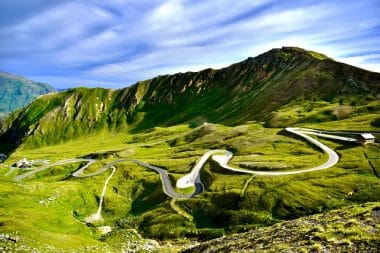Eine kurvenreiche Bergstraße, die als eine der schönsten Panoramastraßen Europas gilt, schlängelt sich unter blauem Himmel mit zarten Wolken durch üppig grüne Hügel, im Hintergrund ragen schroffe Gipfel empor.