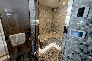 Moderne Dampfkabine mit Mosaikfliesen an den Wänden, einer eingebauten Sitzbank, einer Glastür mit Handtuchhalter und einem Bedienfeld an der Wand. Sanfte Beleuchtung unterstreicht die Spa-ähnliche Atmosphäre.