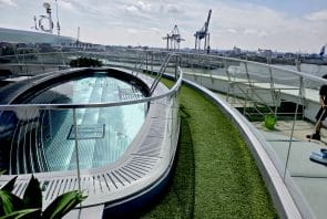 Ein moderner Dachpool mit klarem Wasser liegt neben einem geschwungenen Weg aus Kunstrasen und bietet unter einem teilweise bewölkten Himmel den Blick auf einen Industriehafen mit großen Kränen im Hintergrund.