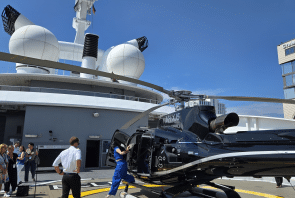 Ein schwarzer Hubschrauber steht auf einem Landeplatz auf dem Deck eines Schiffes, daneben stehen Besatzungsmitglieder. Unter blauem Himmel beobachten Menschen die Szene und machen Fotos, im Hintergrund sind Schiffsstrukturen und ein Gebäude zu sehen.