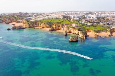 Luftaufnahme der Küstenstadt Lagos an der Algarve mit türkisfarbenem Wasser, felsigen Klippen, Sandstränden und Booten, die weiße Spuren auf dem Meer hinterlassen. Im Hintergrund sind Gebäude und Grünflächen zu sehen, die einige der atemberaubendsten Sehenswürdigkeiten der Region zeigen.