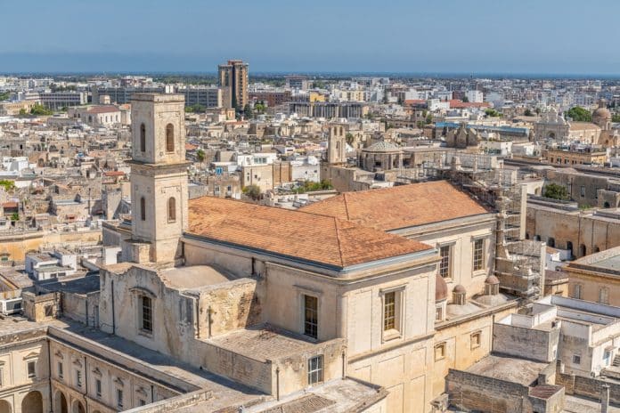 Luftaufnahme einer Stadt mit historischen, beigen Steinhäusern, einem markanten Kirchturm und einer dichten Stadtlandschaft, die sich unter strahlend blauem Himmel bis zum Meer erstreckt – eine der faszinierendsten Sehenswürdigkeiten von Lecce.