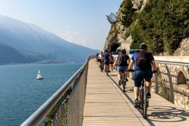 Radfahrer fahren auf einem Holzsteg am Seeufer in Limone sul Garda entlang, im Hintergrund sind Berge zu sehen und auf dem Wasser ein Segelboot. Der Weg verläuft neben einer felsigen Klippe, auf der angrenzenden Straße sind Autos zu sehen.
