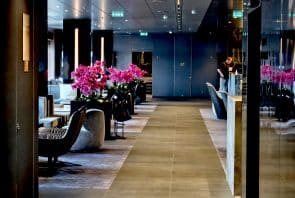 Moderne Hotellobby mit bequemen Sesseln, leuchtend rosa Blumenarrangements auf den Tischen und einer Person, die im Hintergrund neben einer Theke steht. Der Raum ist hell beleuchtet und zeichnet sich durch spiegelnde Oberflächen sowie ein elegantes, modernes Dekor aus.