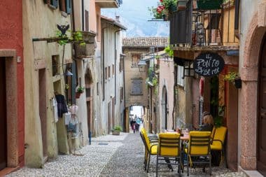 Eine schmale Kopfsteinpflasterstraße in einem europäischen Dorf mit bunten Gebäuden, Café-Tischen im Freien, Pflanzen und Menschen, die in der Ferne spazieren gehen.