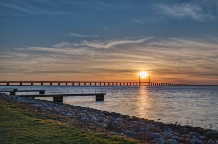 Sunset,On,The,Oresund,Bridge,(oresundsbron),In,The,Baltic,Sea. Eine lange Brücke überspannt bei Sonnenuntergang das ruhige Wasser, während die Sonne tief am Horizont steht und Wolken den Himmel durchziehen. Im Vordergrund sind eine felsige Uferlinie und ein kleiner Steg zu sehen, die eine malerische Szenerie bilden, die an die berühmten Sehenswürdigkeiten von Malmö erinnert.