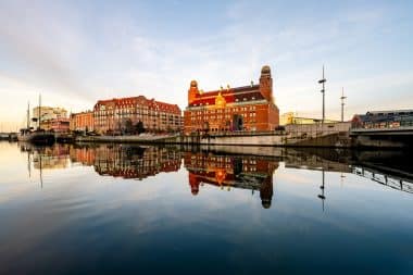 Historische Gebäude aus rotem Backstein und Türme spiegeln sich unter blauem Himmel im ruhigen Wasser wider, im Hintergrund sind Boote und moderne Bauwerke zu sehen – ein Bild, das einige der bekanntesten Sehenswürdigkeiten Malmös einfängt.