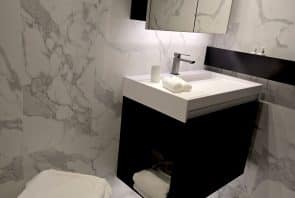 Modernes Badezimmer mit Wänden und Boden aus weißem Marmor, einem schwarz-weißen Waschtisch mit Spiegelschrank und einem weißen Hocker, auf dem ein gefaltetes Handtuch liegt. Auf dem Waschtisch stehen eine Tasse und ein Seifenspender.