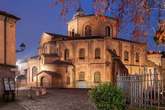 Ravenna,,Italy,At,The,Historic,Basilica,Of,San,Vitale,In Eine alte Backsteinkirche mit Bogenfenstern, die in der Abenddämmerung in warmes Licht getaucht ist. Von einem Baum auf der rechten Seite hängen rote Blätter herab. Im Vordergrund sind ein Kopfsteinpflasterweg und ein Eisenzaun zu sehen.