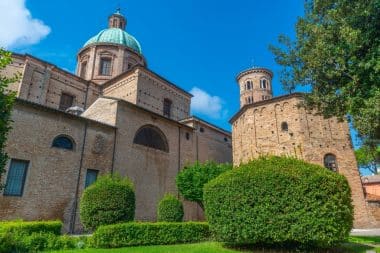 Historische Backsteinkirche mit einer großen grünen Kuppel und einem runden Turm, umgeben von sorgfältig geschnittenen Sträuchern und Bäumen unter strahlend blauem Himmel.