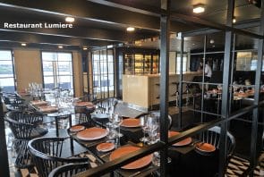 Ein modernes Restaurantinterieur mit schwarzen Stühlen, Tischen, auf denen orangefarbene Teller und Gläser stehen, sowie einer Spiegelwand. Links ist das Schild „Restaurant Lumiere“ zu sehen. Im Hintergrund sind ein Barbereich und große Fenster zu erkennen.