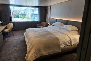 Modernes Hotelzimmer mit einem großen, ordentlich gemachten Bett, einem Schreibtisch, einem Stuhl, einem Sessel und einem großen Fenster, das einen Blick auf die Stadt bietet, wobei draußen Gebäude und Bäume zu sehen sind.