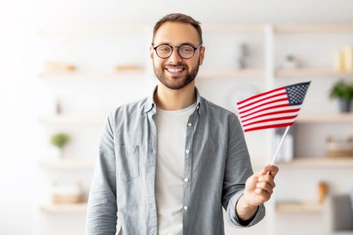 Handsome,Young,Guy,Holding,American,Flag,And,Smiling,At,Camera Ein lächelnder Mann mit Brille hält in einem Raum eine kleine amerikanische Flagge in der Hand und steht vor verschwommenen Regalen.