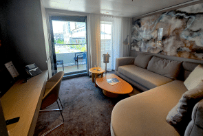 Modernes Hotelzimmer mit einem Sofa, einem Schreibtisch und zwei kleinen Tischen. Das raumhohe Fenster führt auf einen Balkon mit Stühlen. Das Licht von draußen erhellt den Raum, und an einer Wand hängt ein abstraktes Kunstwerk.